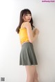 A woman in a yellow top and green skirt leaning against a wall.